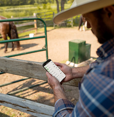 A man in a cowboy hat and plaid shirt uses the Polaris RIDE COMMAND app on a smartphone while standing by a wooden fence, with horses in the background on a ranch.