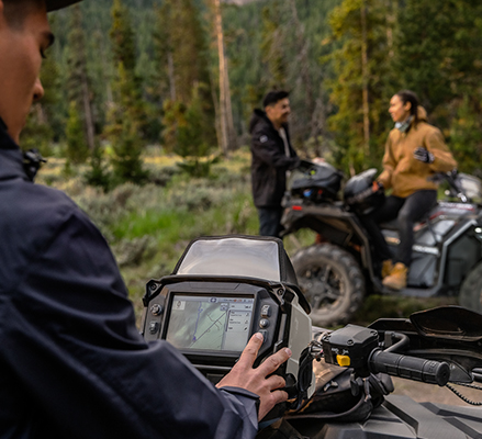 A person operating a 2025 Polaris Sportsman 570 ATV with the RIDE COMMAND digital touch screen display on the handlebar showing a Group Ride GPS map, with a couple with another 2025 Polaris Sportsman 570 ATV in the background on a forested trail.