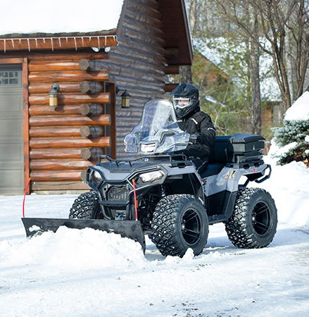 Person operating a black 2025 Polaris Sportsman 570 ATV with a snow plow attachment, clearing snow in front of a cabin-style house in winter.