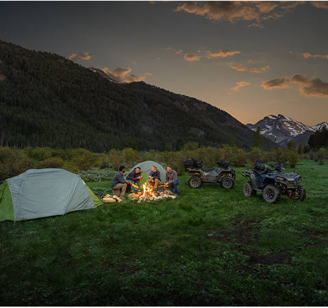 4 people sitting around a campfire, near tents that were hauled with their 2025 Polaris Sportsman Touring 570 2-up ATVs.