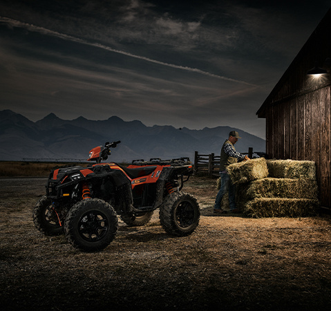 Side profile of a 2025 Polaris Sportsman XP 1000 S ATV with a man stacking hay bales near a barn in the background.