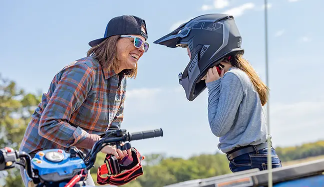 A parent smiling at her child while she puts on her helmet before a ride on a 2026 Polaris Sportsman 110 youth ATV.