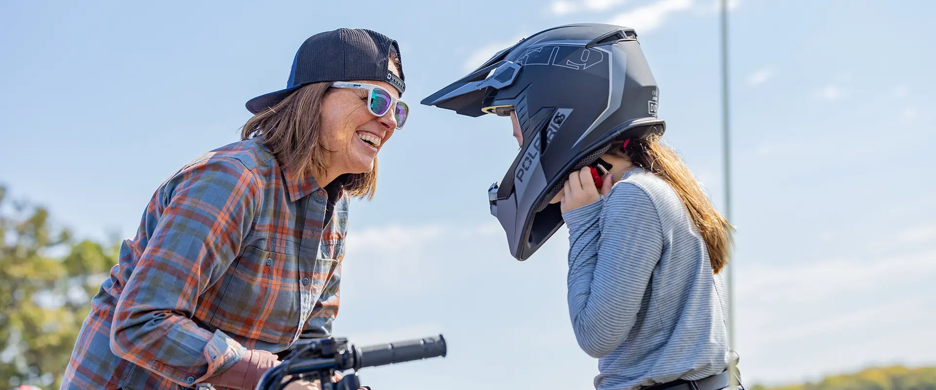 A parent smiling at her child while she puts on her helmet before a ride on a 2026 Polaris Sportsman 110 youth ATV.