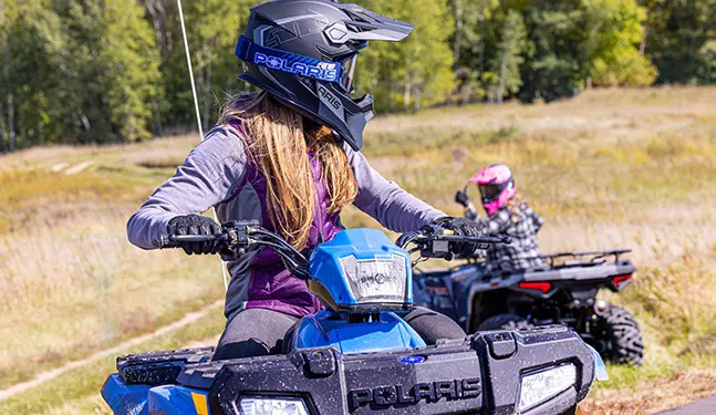 A girl with a helmet on driving a 2026 Polaris Sportsman 110 youth 4-wheeler and looking back toward another kid on an quad.