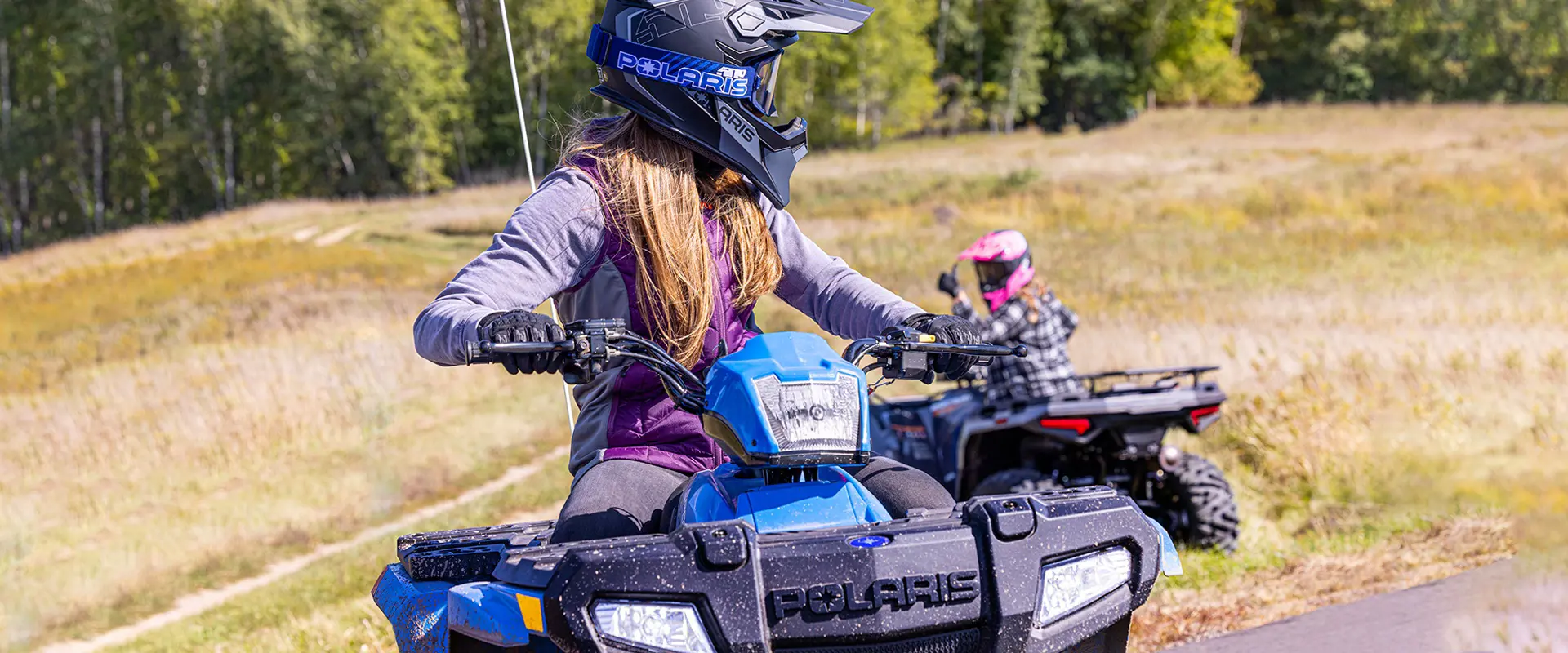 A girl with a helmet on driving a 2026 Polaris Sportsman 110 youth 4-wheeler and looking back toward another kid on an quad.