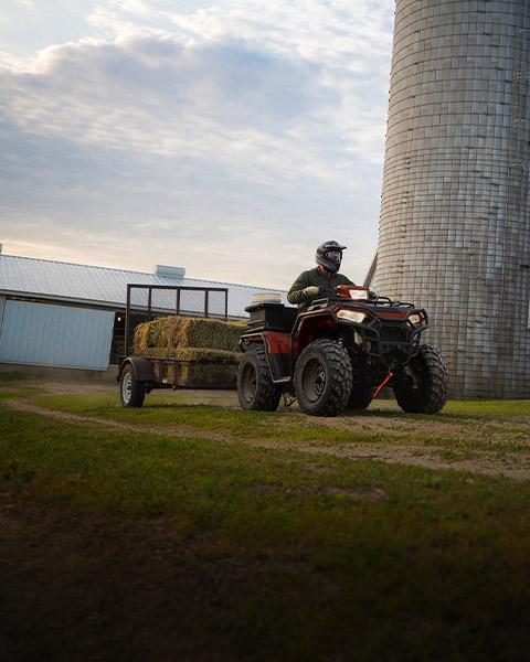 A Polaris Sportsman 450 ATV pulls hay bales in a grassy field near a rustic barn.