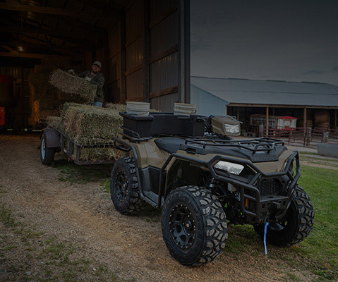 Polaris Sportsman ATV with front and rear storage accessories parked on a farm, in front of a hay-loaded trailer and surrounded by barns and agricultural equipment.
