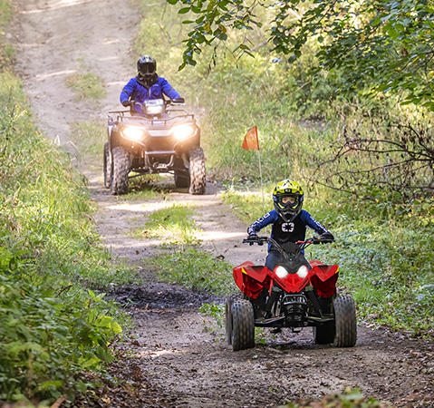 A riding an Indy Red 2027 Polaris Outlaw 110 EFI youth ATV while his parent drives behind on a trail.