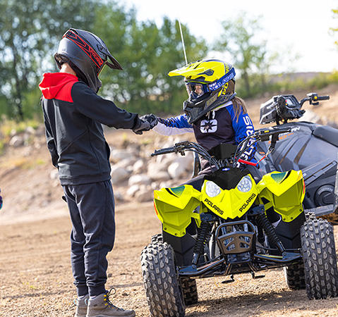 A kid giving knuckles from the seat of their 2027 Polaris Outlaw 110 EFI youth ATV in the Lime Squeeze colorway.