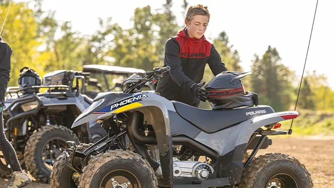 A boy grabbing his helmet to ride his 2027 Polaris Phoenix 200 youth ATV.