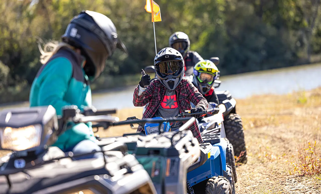 A kid giving a thumbs up from a 2027 Polaris Sportsman 110 EFI youth ATV to other youth ATV riders.