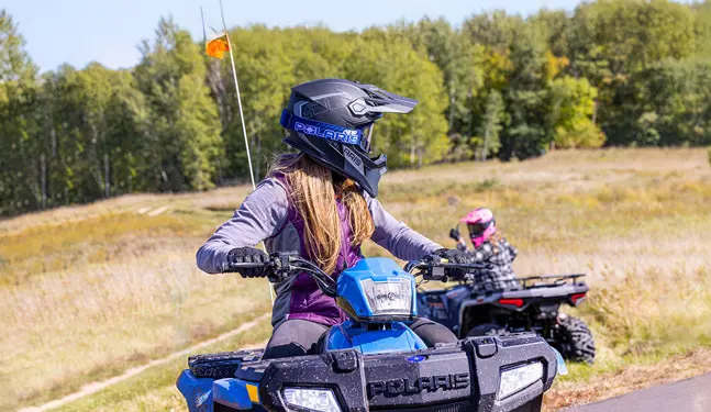 A rider on a 2027 Polaris Sportsman 110 EFI youth ATV looking back at another rider on a Polaris youth ATV.
