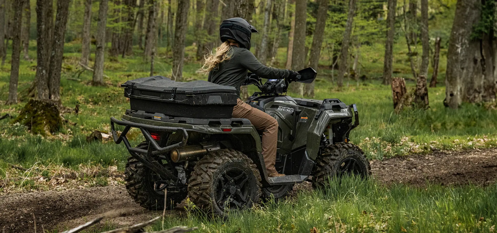 Rear view of a woman on a 2027 Polaris Sportsman 500 ATV cruising down a forest trail.