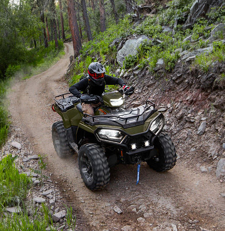 A rider on a 2027 Polaris Sportsman 600 ATV with LED pod and headlights illuminated riding on a trail.