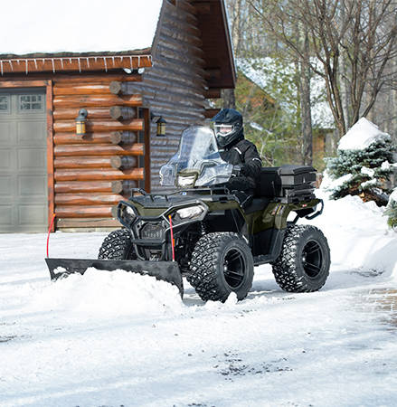A person on a 2027 Polaris Sportsman 600 ATV using a plow to move snow in the winter.
