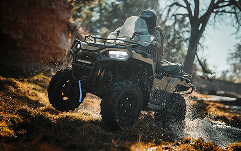 Polaris Sportsman ATV crossing a shallow stream in a wooded landscape, splashing water while navigating rugged terrain with a rider and cargo mounted on the rear rack.