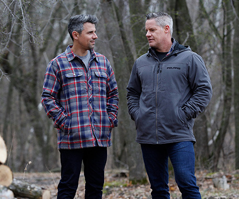 two men chatting in the woods, wearing Polaris apparel, one of them wearing a flannel jacket