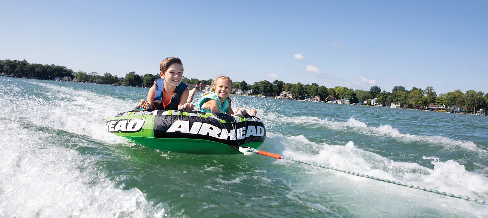 Two kids smiling while tubing behind a Bennington RX Sport pontoon boat.