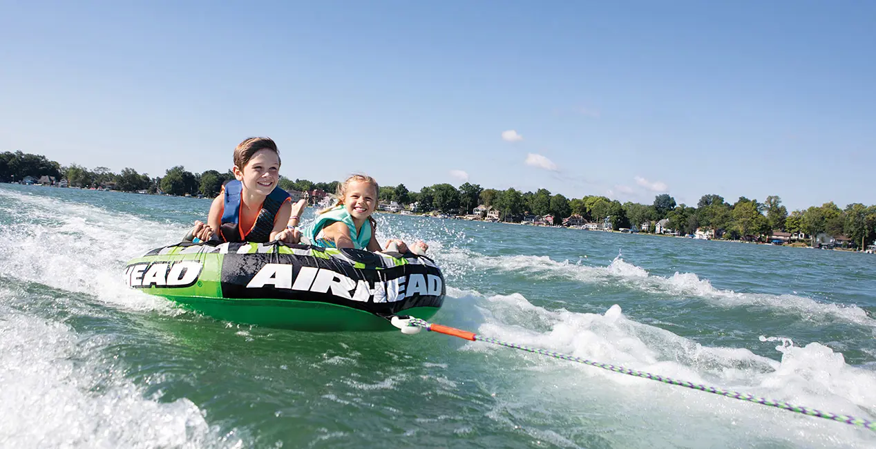 Two kids smiling while tubing behind a Bennington RX Sport pontoon boat.