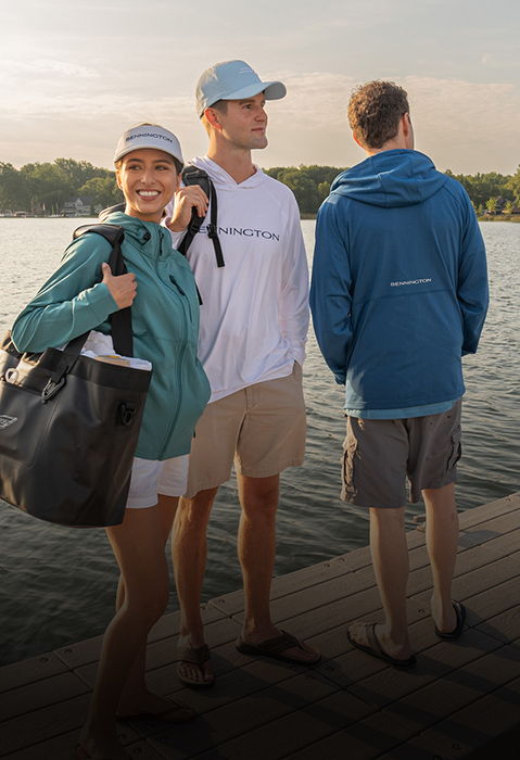 Three young adults on a dock by the lake, wearing Bennington branded apparel including a white long-sleeve shirt, blue hoodie, and visor.
