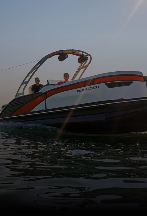 A Bennington Pontoon Boat pulls a waterskier across a lake at sunset, with two people visible on the boat.