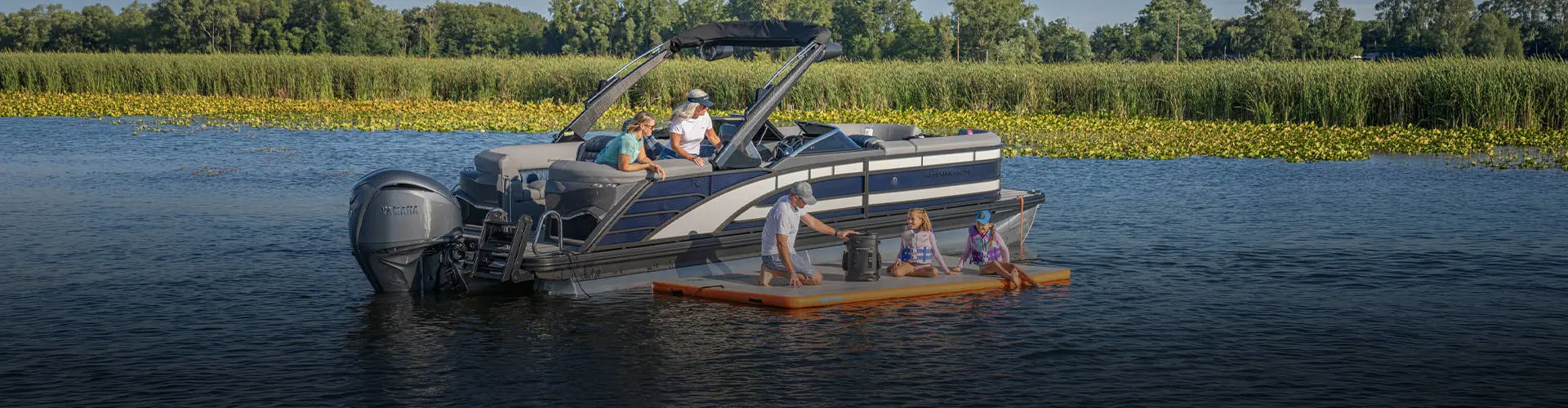 Man unzips backpack on a floating lily pad with family next to a pontoon anchored on the lake. 