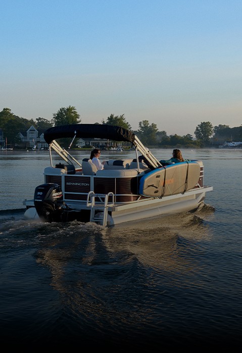 Two women passengers cruise on a Bennington pontoon boat carrying a paddleboard across a calm lake.