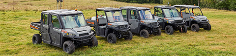 A line of five grey and orange Polaris Commercial Pro XD UTVs are parked in a grassy field with trees in the background.