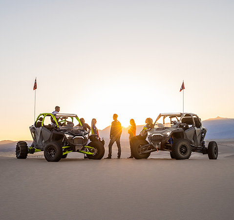 A group of Polaris Off-Road ambassadors & partners with next to their RZR vehicles at sunset in desert, silhouetted against a bright sky.