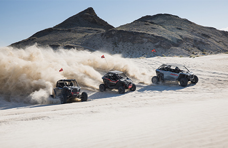3 rzr riding uphill on the desert dunes, dust flying all arround