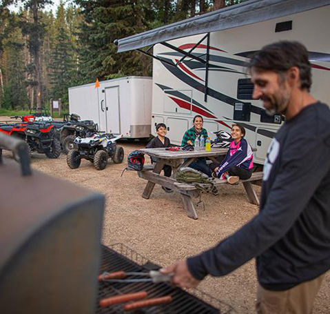 Familia cocinando a la parrilla en una mesa de pícnic con un vehículo recreativo y ATV Polaris Sportsman estacionados cerca en un campamento.