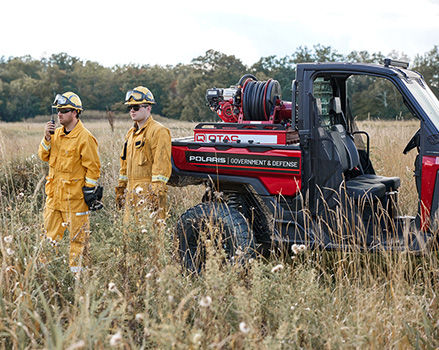 Two firefighters in yellow gear stand near a red Polaris Government and Defense UTV equipped with a water tank and hose reel, in a grassy field.