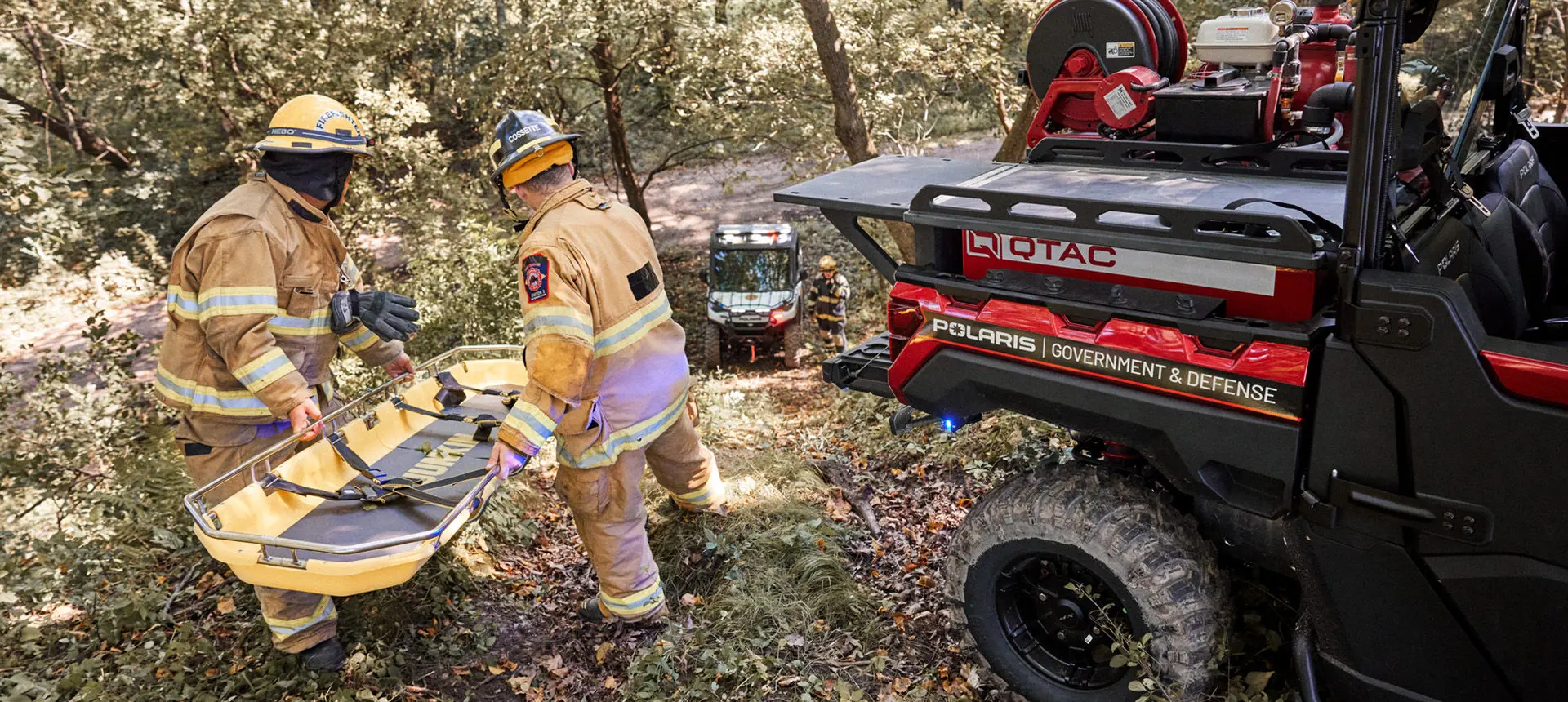 Two firefighters in turnout gear carry a yellow rescue basket stretcher through a wooded area towards a red Polaris Government & Defense UTV. Another UTV and firefighter are visible in the background.