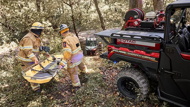 Two firefighters in turnout gear carry a yellow rescue basket stretcher through a wooded area towards a red Polaris Government & Defense UTV. Another UTV and firefighter are visible in the background.