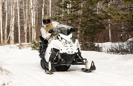 A person wearing winter camouflage gear rides a white and black snowmobile 2026 Military 850 TITAN 155 on a snowy path lined with trees.