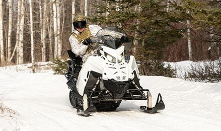 A person wearing winter camouflage gear rides a white and black snowmobile 2026 Military 850 TITAN 155 on a snowy path lined with trees.