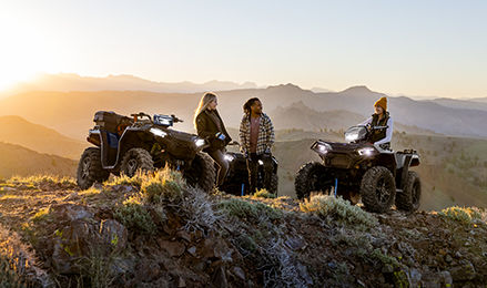 Three riders with Polaris ATVs enjoying a sunset view on a mountain trail.