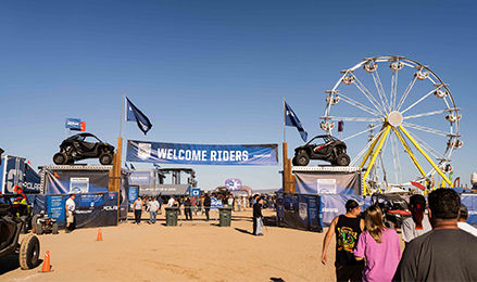 Welcome banner and Polaris RZRs at an outdoor event with a Ferris wheel and crowd.