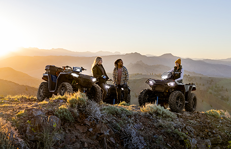 Three riders with Polaris ATVs enjoying a sunset view on a mountain trail.