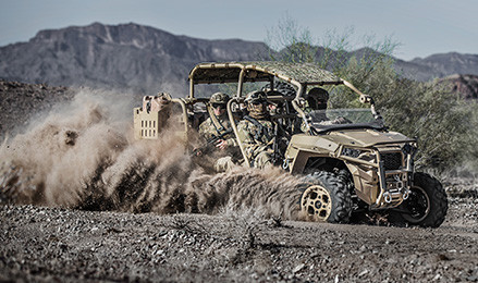 A tan MRZR Diesel light tactical vehicle with four military personnel kicks up a large cloud of dust while driving through a arid, rocky landscape.