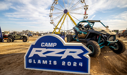 Polaris RZR off-road vehicle beside Camp RZR Glamis 2024 sign, with Ferris wheel and desert event in the background.