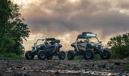 Two Polaris RZR side-by-side vehicles, a Polaris RZR XP and an All New RZR Pro XP S, are parked on a muddy trail at dusk, with trees and a cloudy sky in the background.
