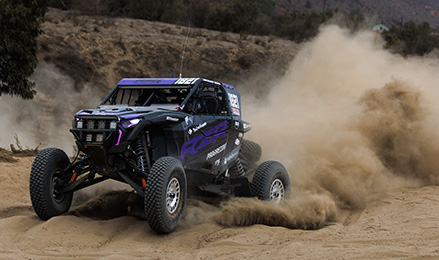 A black and purple Polaris RZR Pro R Factory SxS racing through a dusty desert landscape, kicking up a large cloud of dirt.