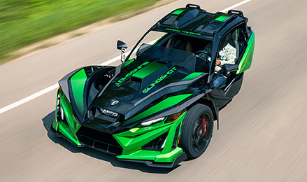 A green and black Polaris Slingshot Grand Touring roadster with a driver is speeding down a paved road, with blurred green scenery in the background.
