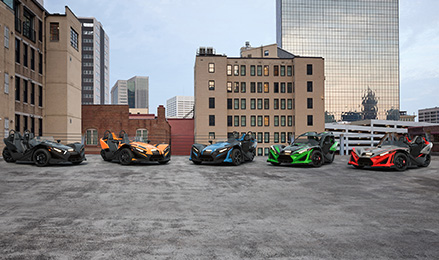 Five colorful Polaris Slingshot vehicles are parked in a line on a rooftop in a city, with buildings and a skyscraper in the background.