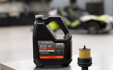 Polaris Slingshot SS-4 Full Synthetic 5W-50 engine oil and oil filter on a metallic surface, with a blurred Polaris Slingshot vehicle in the background.
