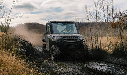 An Avalanche Gray Polaris 2027 RANGER XP 1000 CAB splashes through a muddy trail with a helmeted driver visible inside, surrounded by sparse trees.