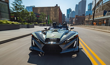 Un Polaris Slingshot Signature Edition de 2026 con dos motociclistas cruzando el puente de una ciudad con un horizonte urbano de fondo.