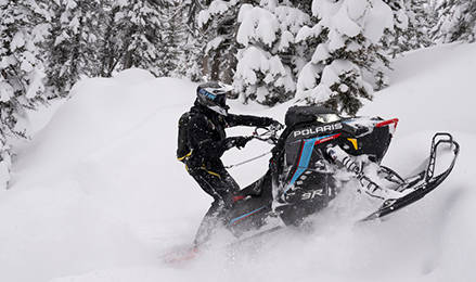 A rider in black gear carves through deep powder on a black and blue Polaris snowmobile RMK Factory Edition in a snow-covered pine forest.
