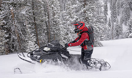 A rider in a red jacket maneuvers a black Polaris INDY VR1 snowmobile through a dense, snow-laden evergreen forest.